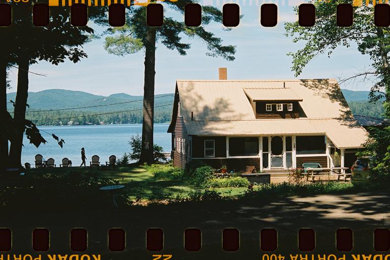 A cottage with a porch sits by a lake with mountains in the background, and a person walks along the shore.