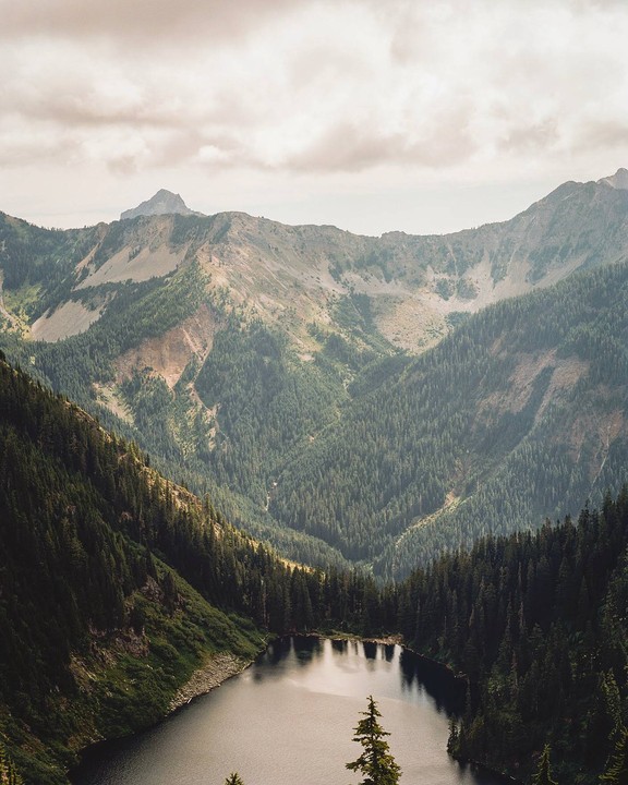 A lake is nestled in a valley surrounded by mountains covered in evergreen trees under a cloudy sky.