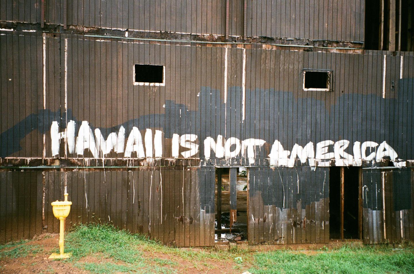 White graffiti on a dark wooden wall reads "HAWAII IS NOT AMERICA" above a yellow fire hydrant and green grass.
