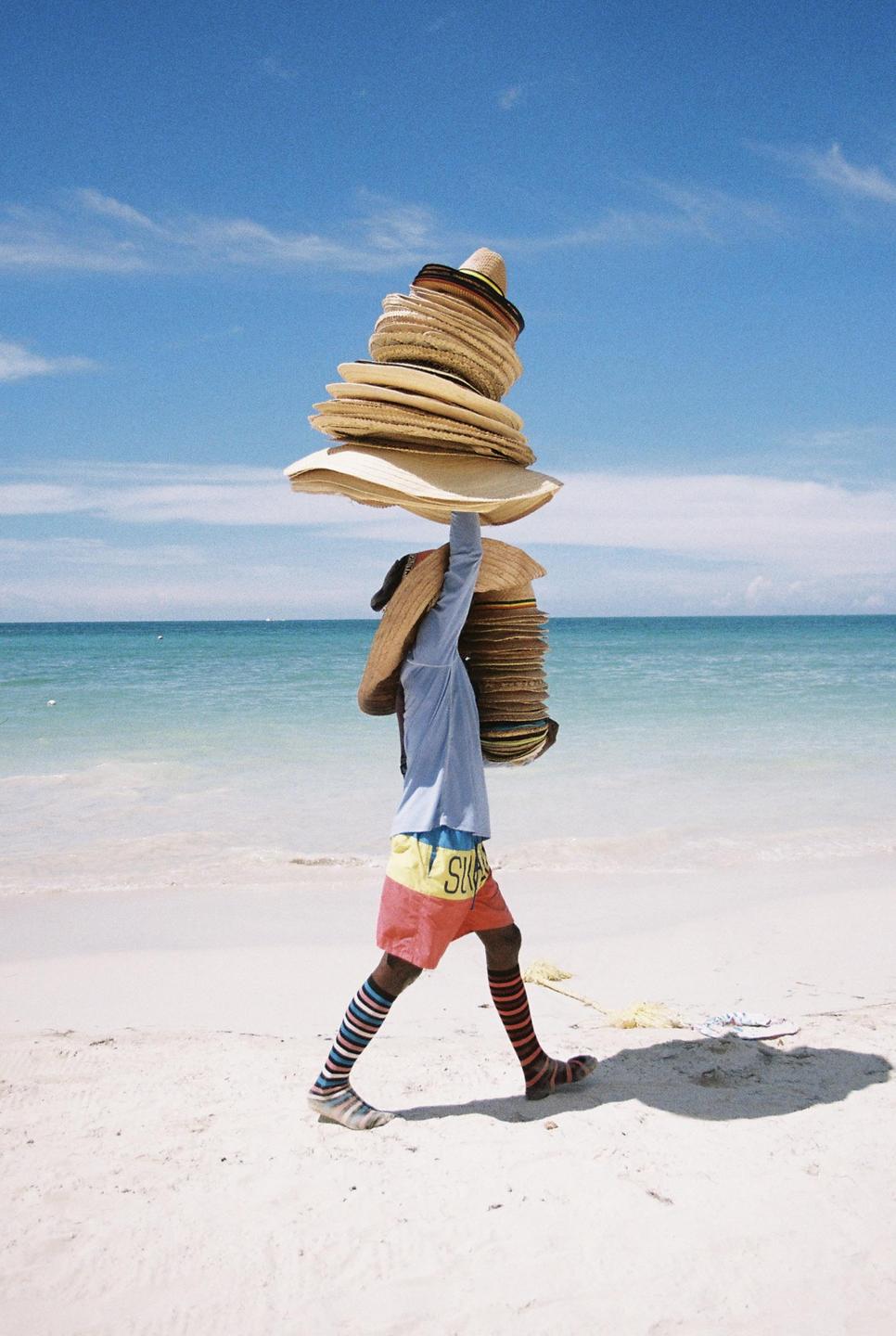 A man walks along a beach carrying a tall stack of hats on his head and another stack in his arm.