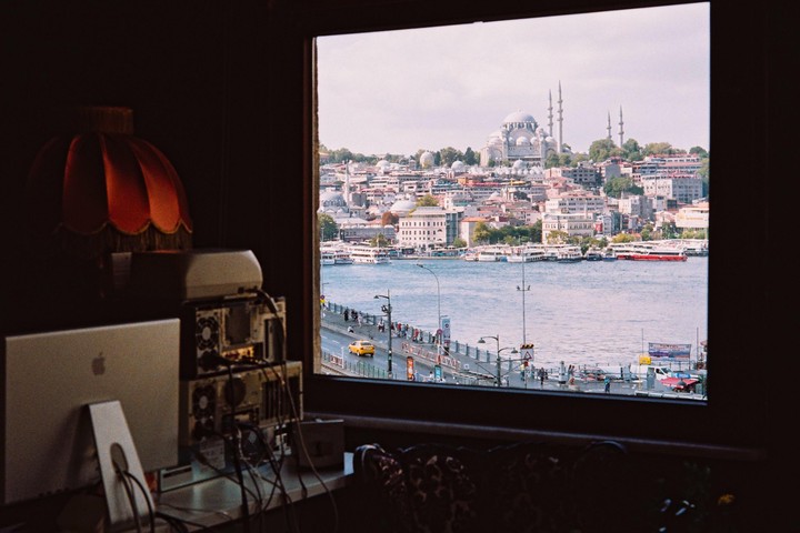 A view of a city with a large mosque and a body of water with boats, seen through a window with a lamp and computer equipment in the foreground.