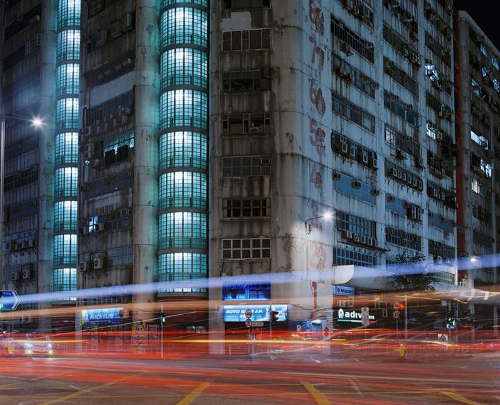 A tall building with many windows and light trails from vehicles on the street below is visible at night.