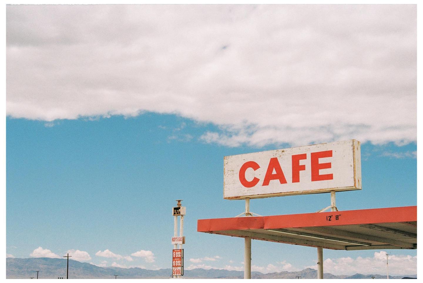 A "CAFE" sign with red lettering is visible above a red awning, with a "ROY'S GAS" sign and mountains in the distance under a blue sky with clouds.