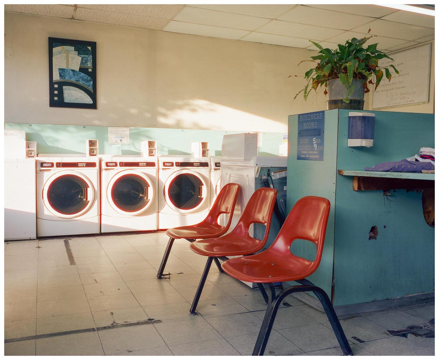Three red chairs are lined up in front of several washing machines in a laundromat.