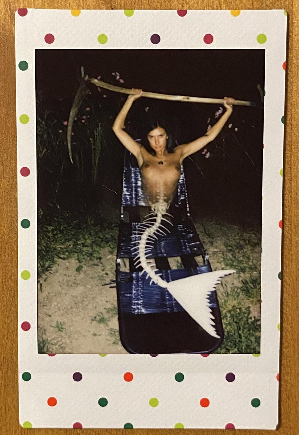 A woman with a fish skeleton tail holds a scythe above her head in a night setting.