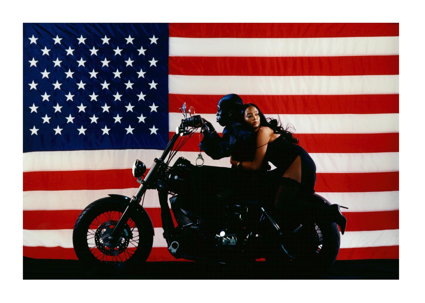A woman in a black dress and a person in a black outfit are on a motorcycle in front of an American flag.