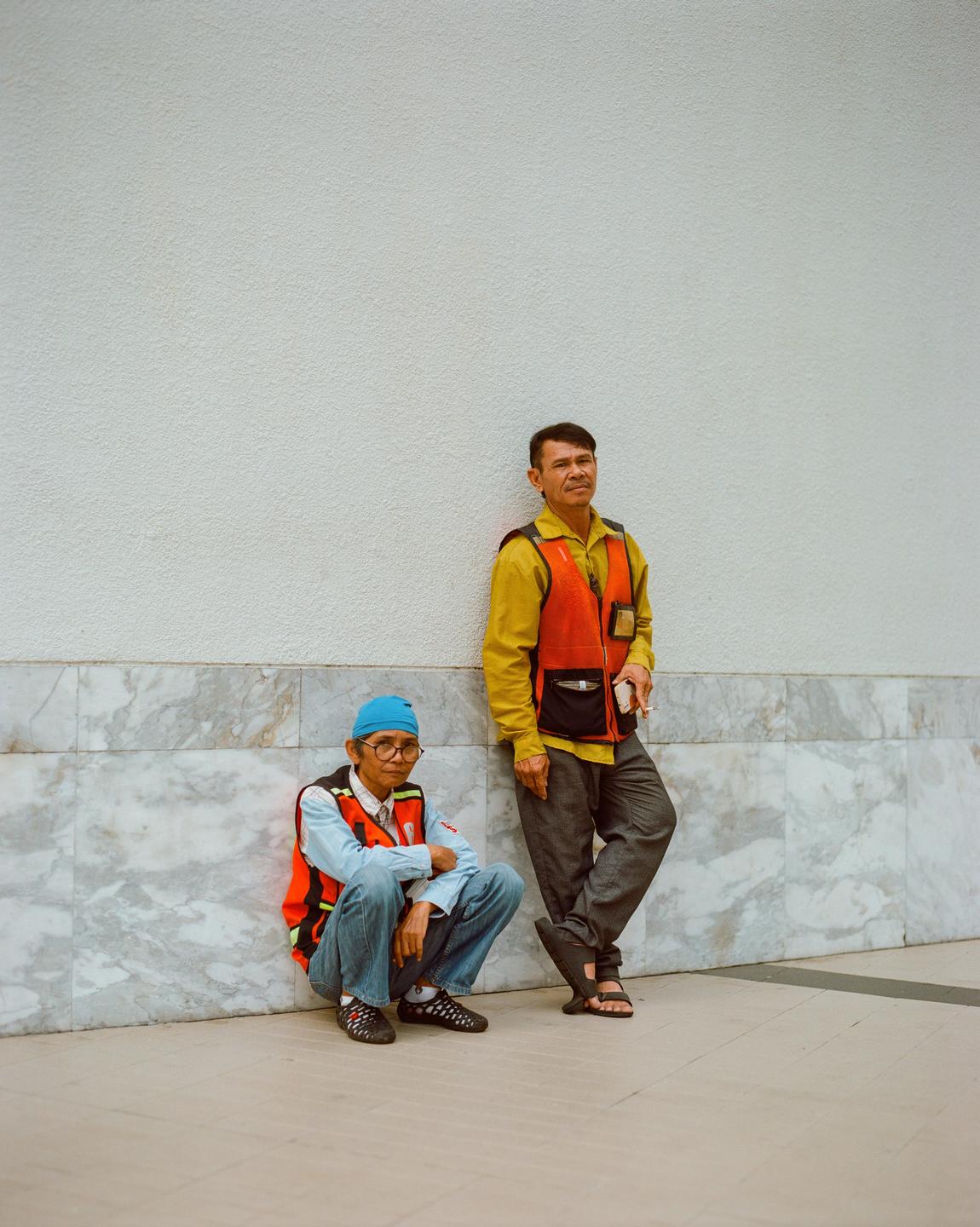 Two men wearing orange safety vests are standing and crouching against a white and marble wall.