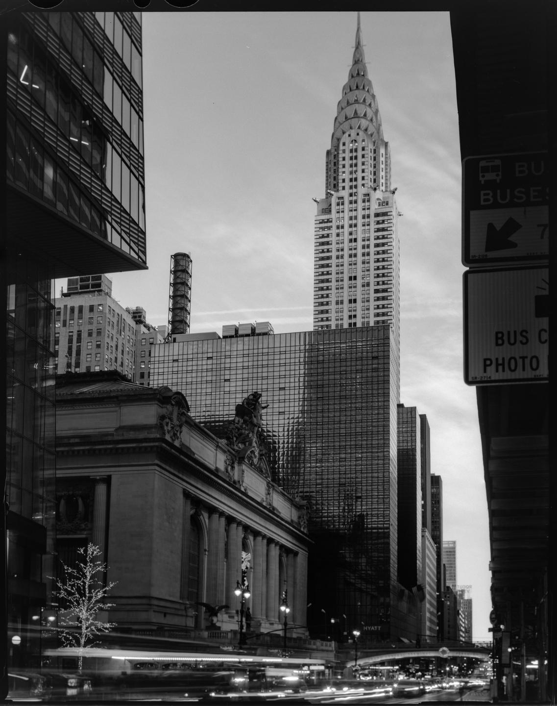 The Chrysler Building rises above Grand Central Terminal and other buildings at a street corner with light trails from traffic.