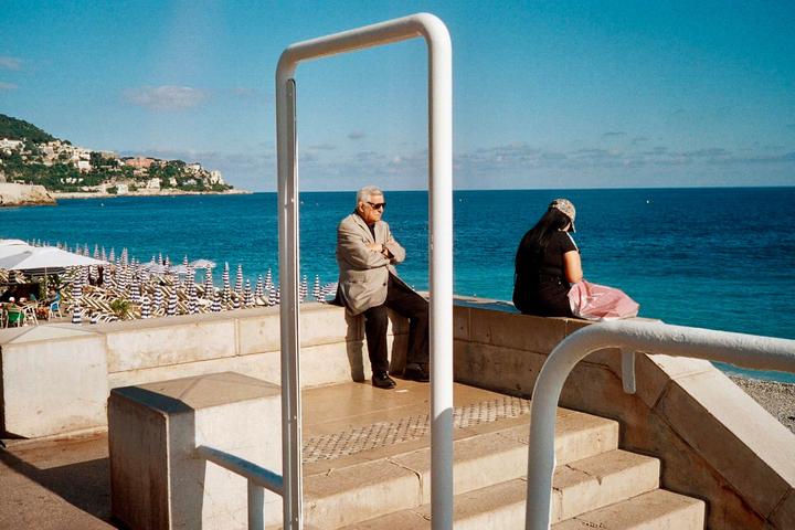 A man and a woman sit on a stone wall overlooking a beach with striped umbrellas and the ocean.