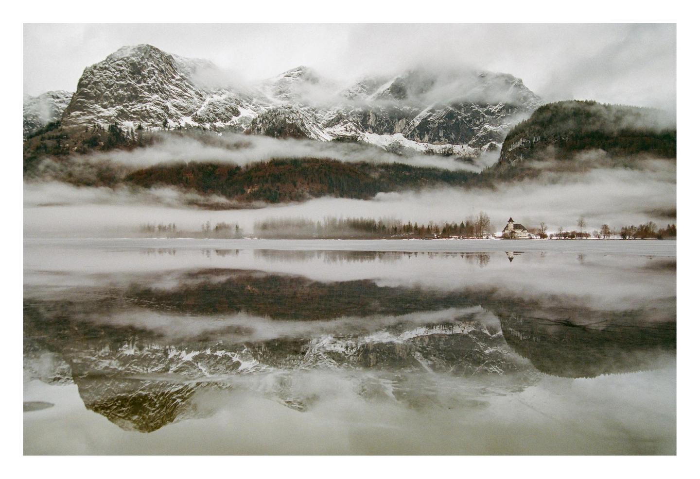 A church and trees are reflected in a frozen lake with snow-capped mountains and fog in the background.