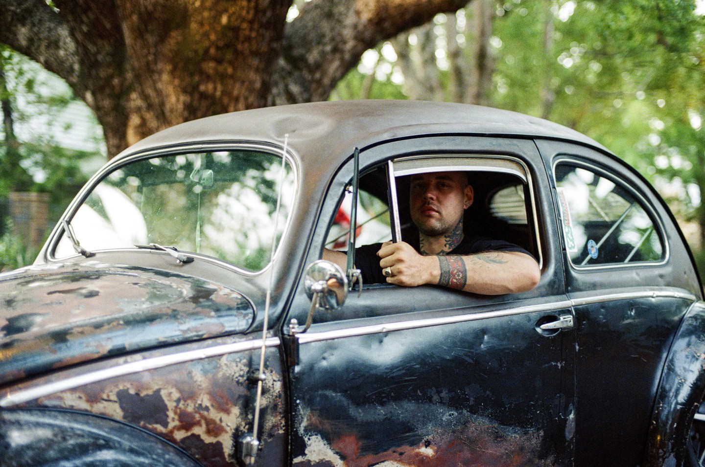 A man with tattoos on his neck and arm sits in a weathered car with his arm resting on the open window.