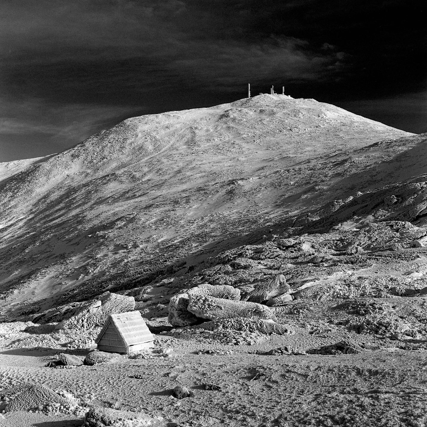 A small A-frame structure sits in a snowy, rocky landscape with a mountain peak topped with antennas in the background.