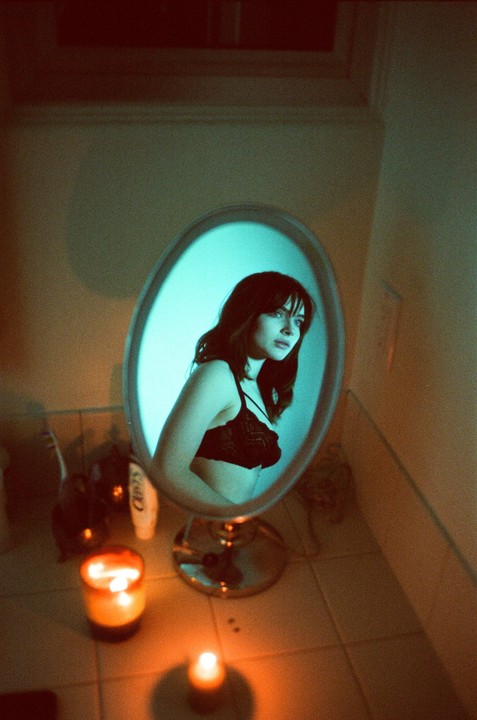 A woman in a black bra is reflected in an oval mirror, illuminated by a blue light, with candles providing warm light in the foreground.
