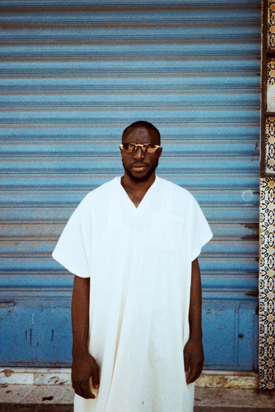 A man wearing a white tunic and sunglasses stands in front of a blue corrugated metal door next to a patterned tile column.