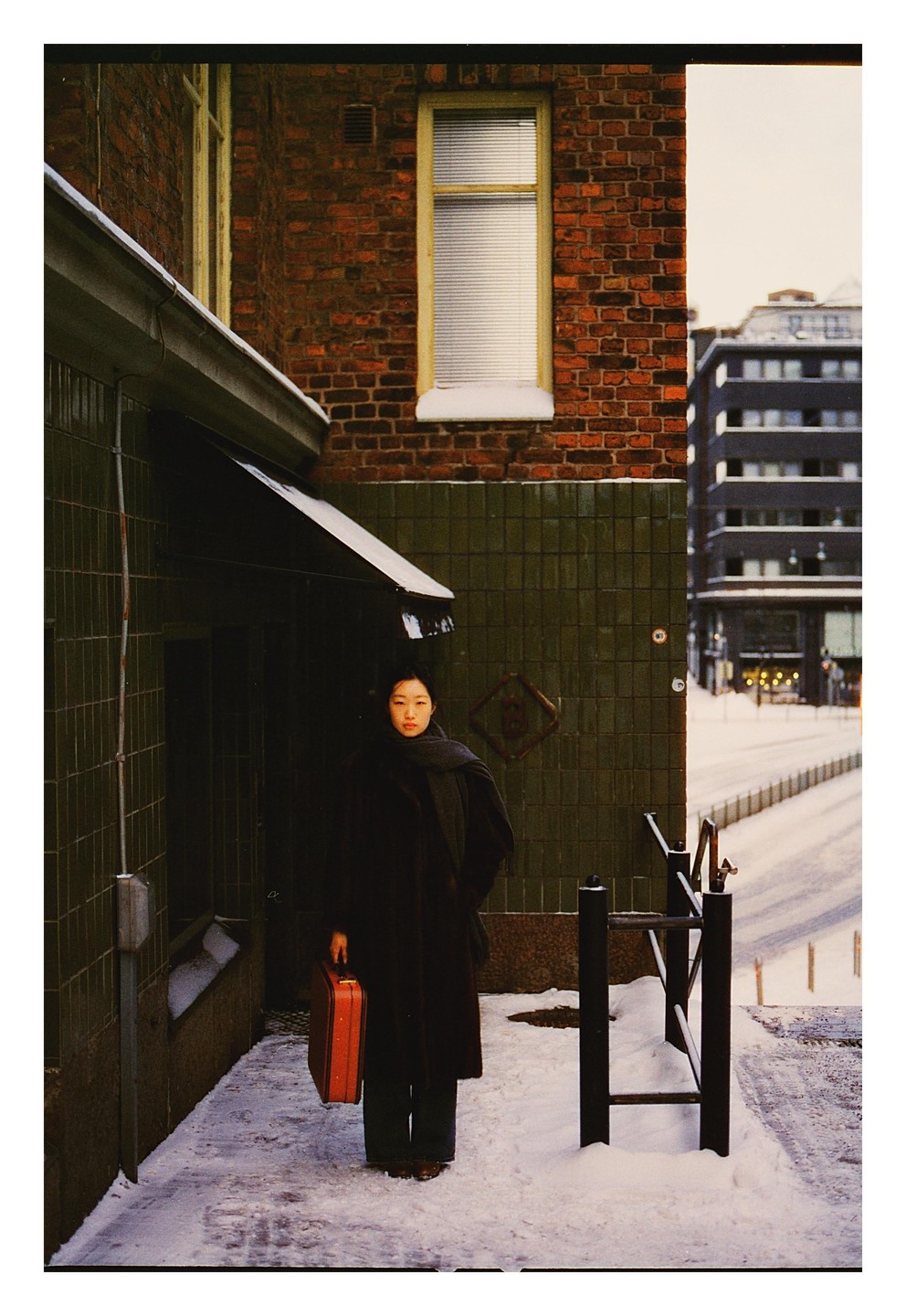 A woman in a dark coat stands on a snowy street next to a brick building, holding a red briefcase.