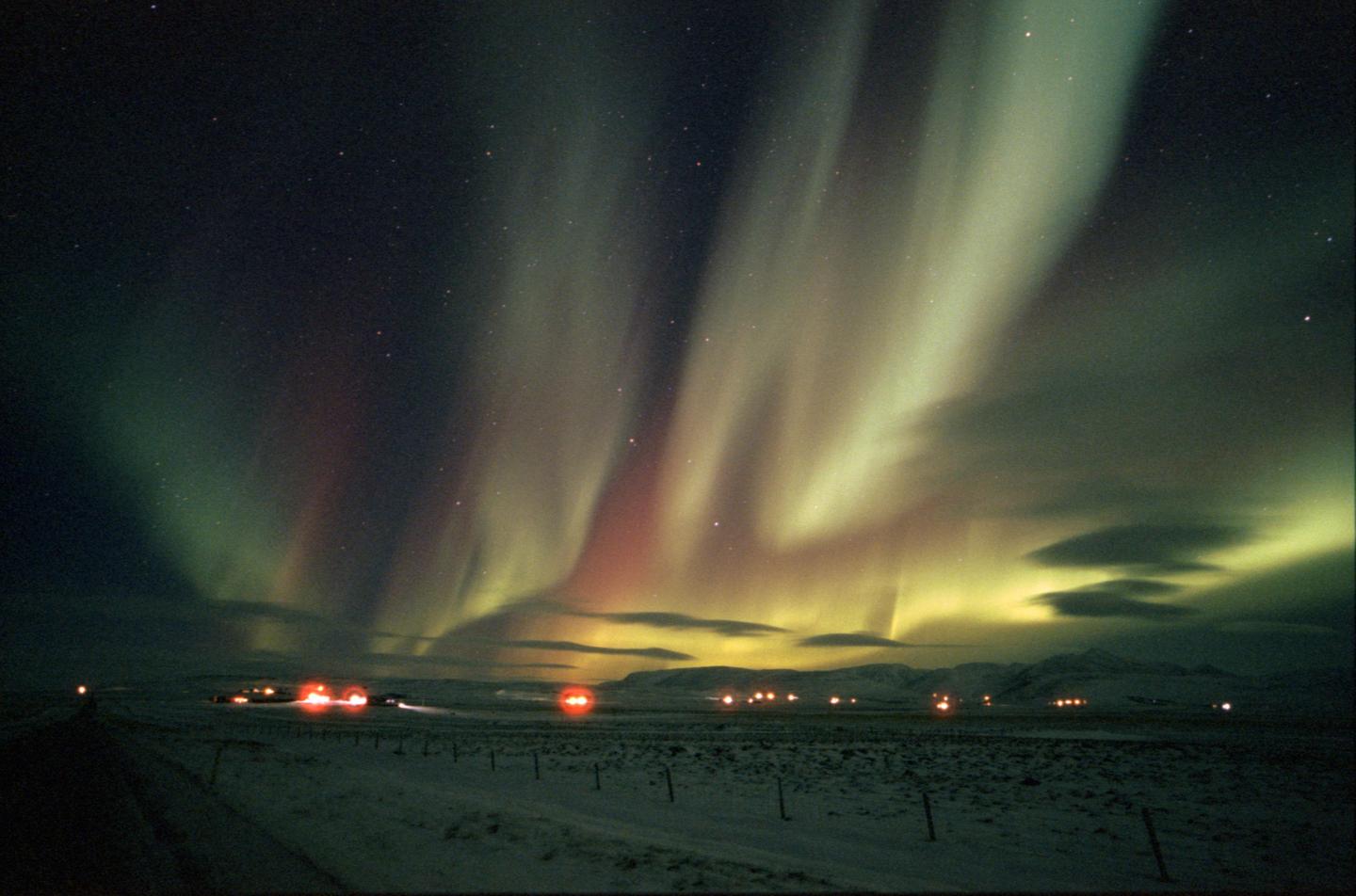 The aurora borealis illuminates a snowy landscape with distant lights and mountains under a starry night sky.