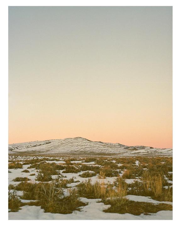 A snow-dusted field with sparse vegetation stretches towards a snow-covered mountain under a sunset sky.