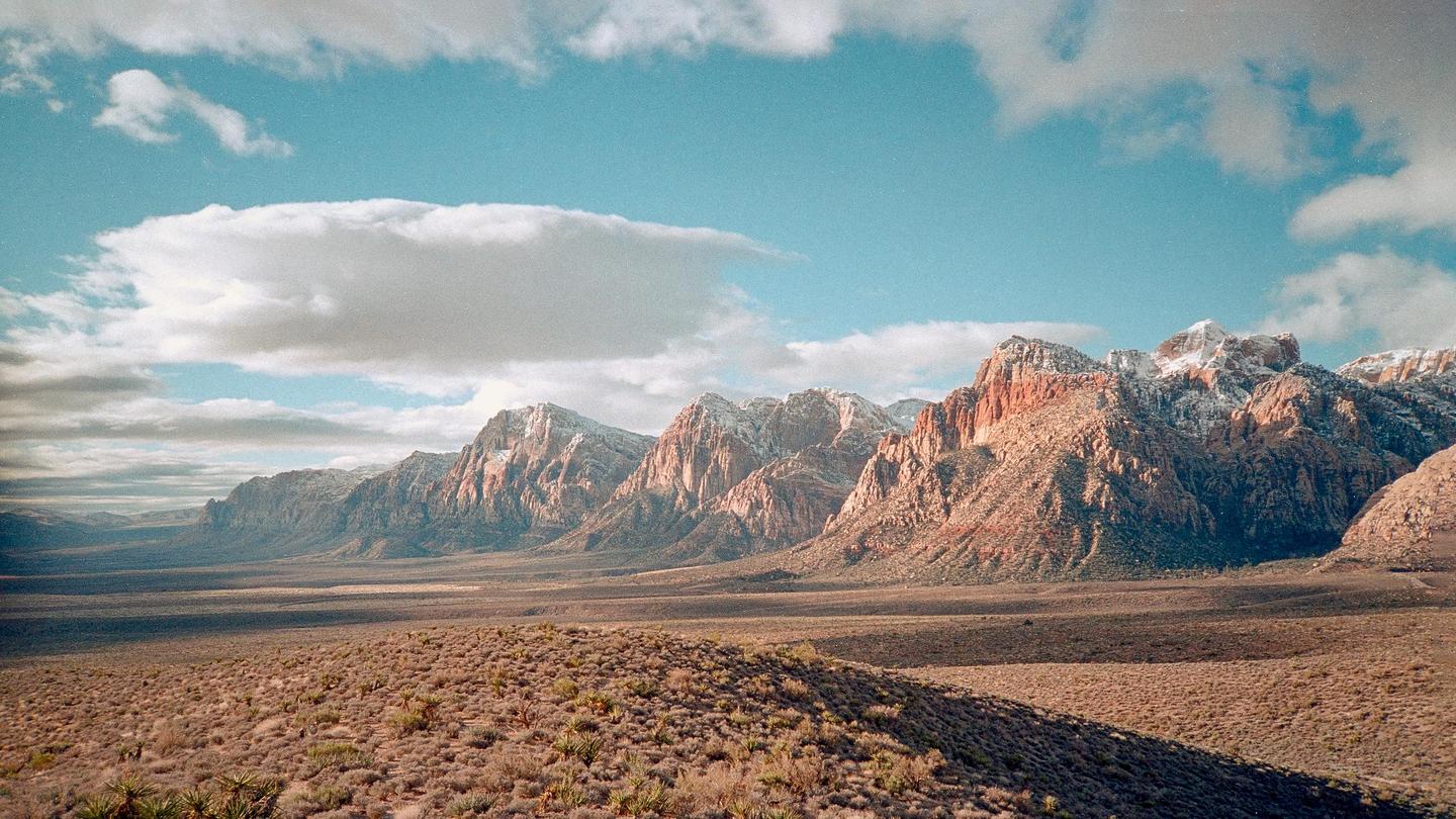 A vast desert landscape with mountains partially covered in snow under a blue sky with clouds.