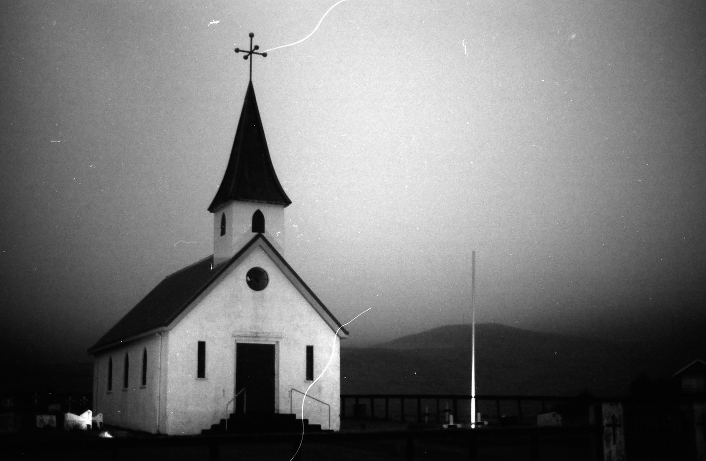 A small white church with a tall steeple stands against a dark, hazy sky with mountains in the distance.