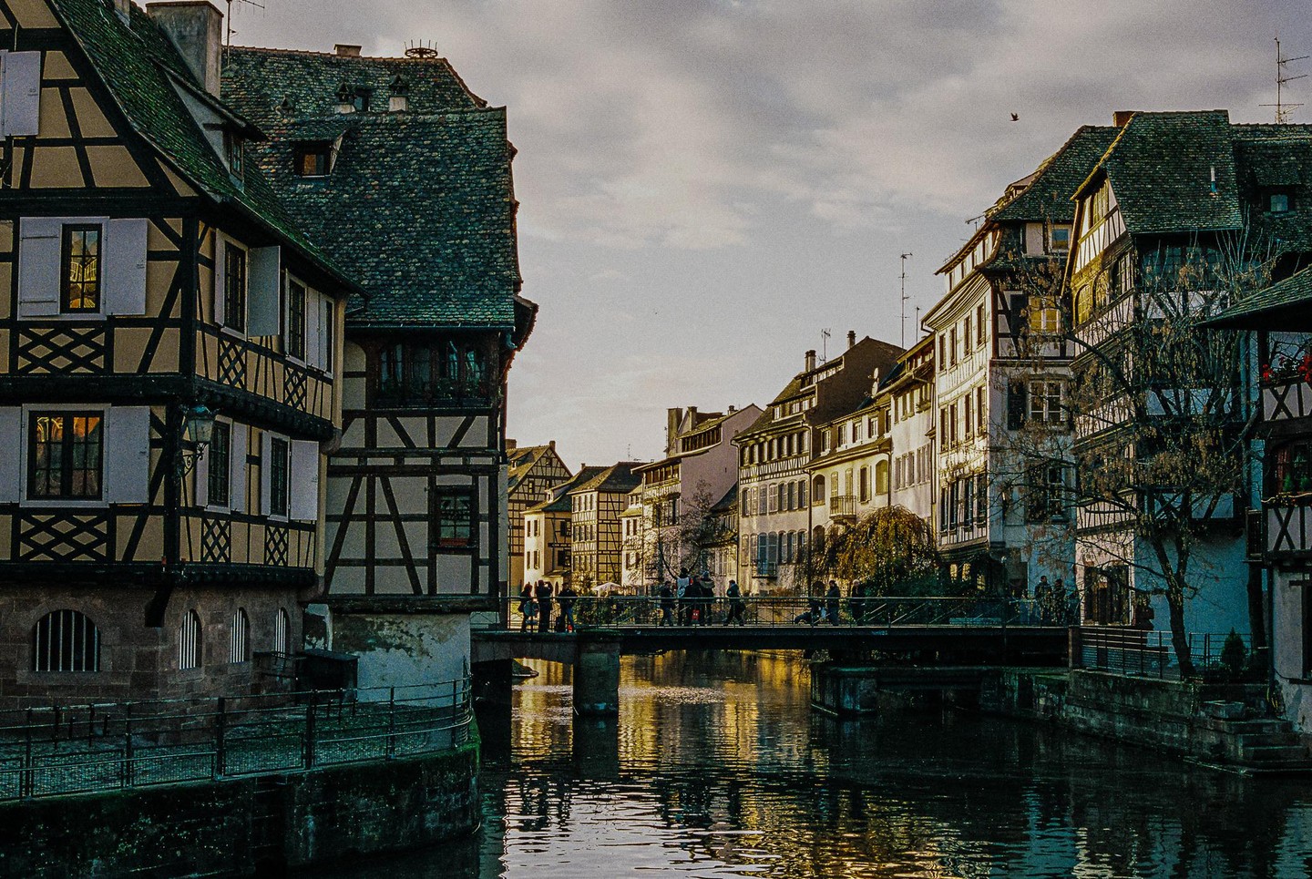 A canal flows through a city with traditional half-timbered buildings and a bridge with people walking across it.