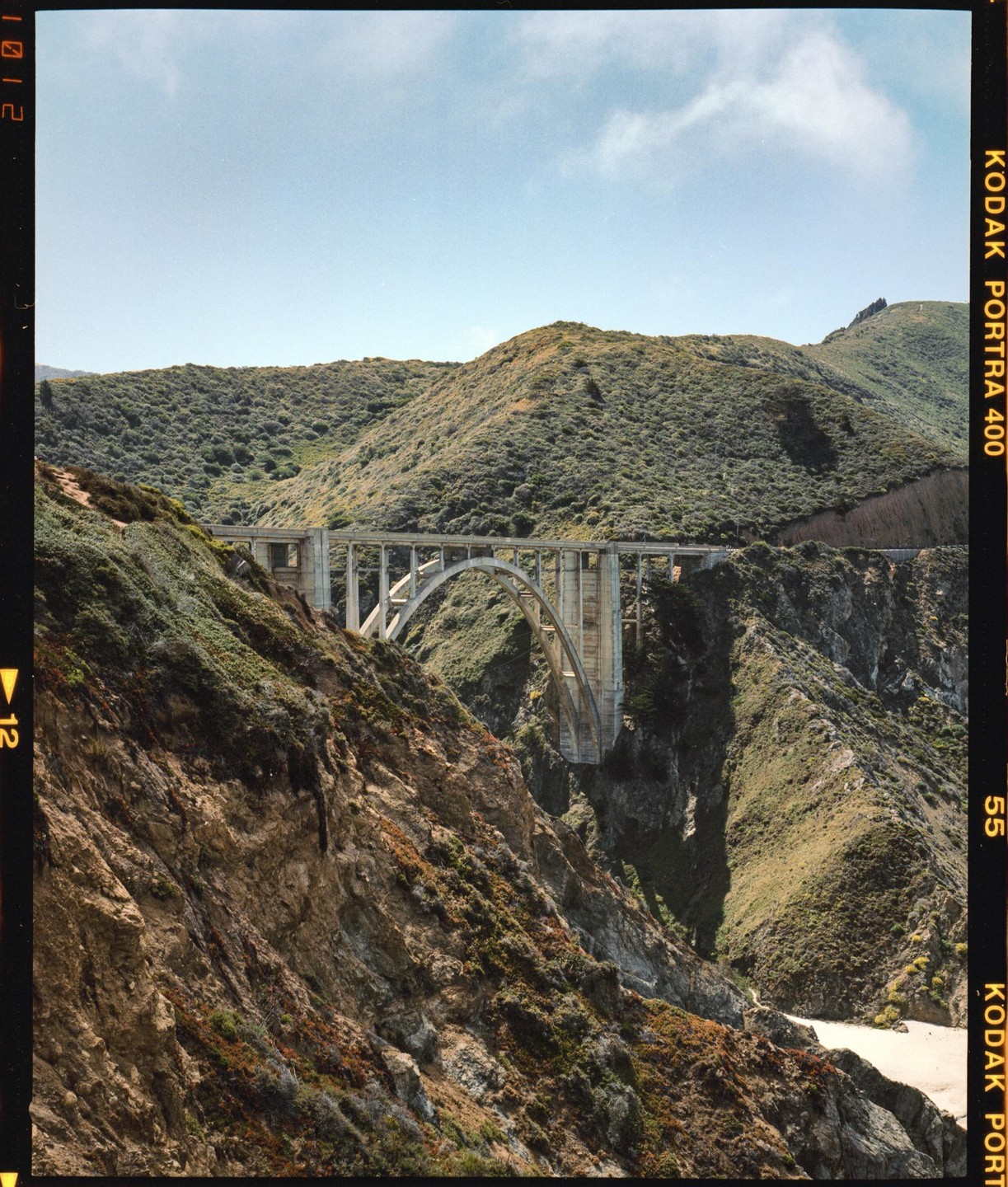 A concrete arch bridge spans a deep canyon with steep, vegetated slopes under a clear sky.