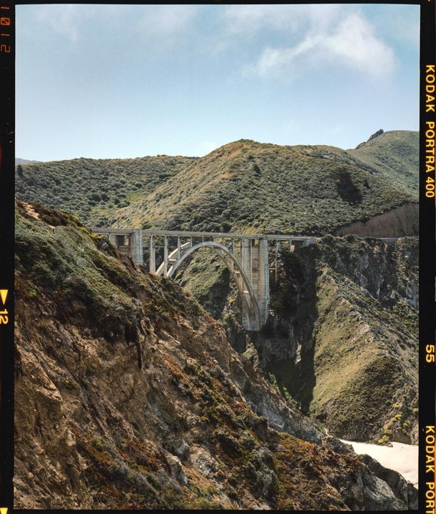 A concrete arch bridge spans a deep canyon with steep, vegetated slopes under a clear sky.