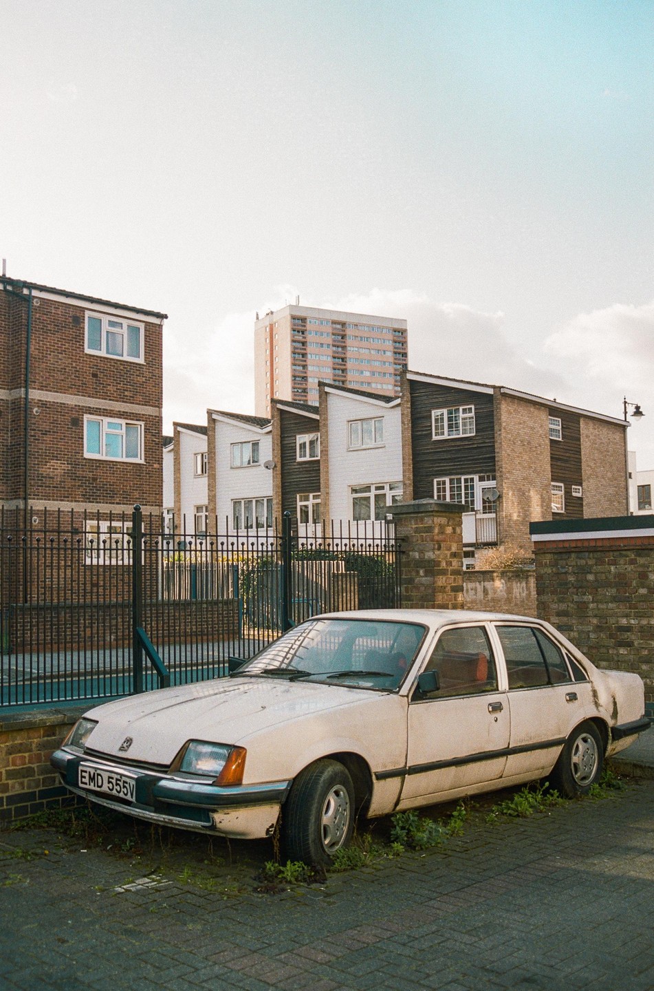A white car is parked on a paved area with grass growing around it, in front of a fence and residential buildings.
