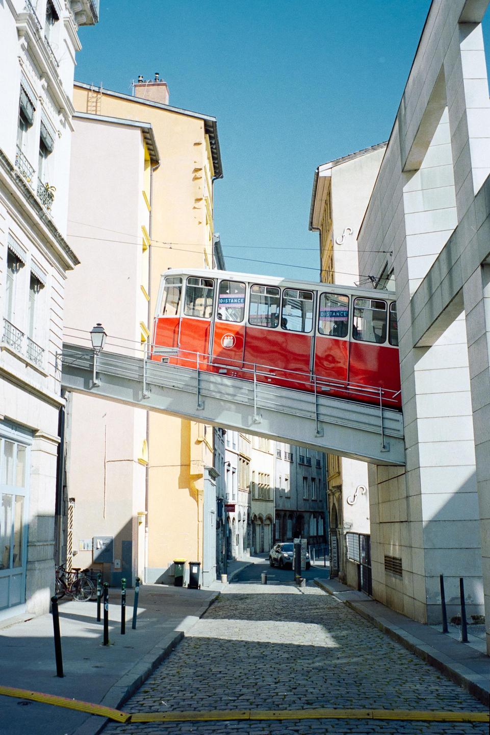 A red funicular car travels on an elevated track between buildings on a sunny day.