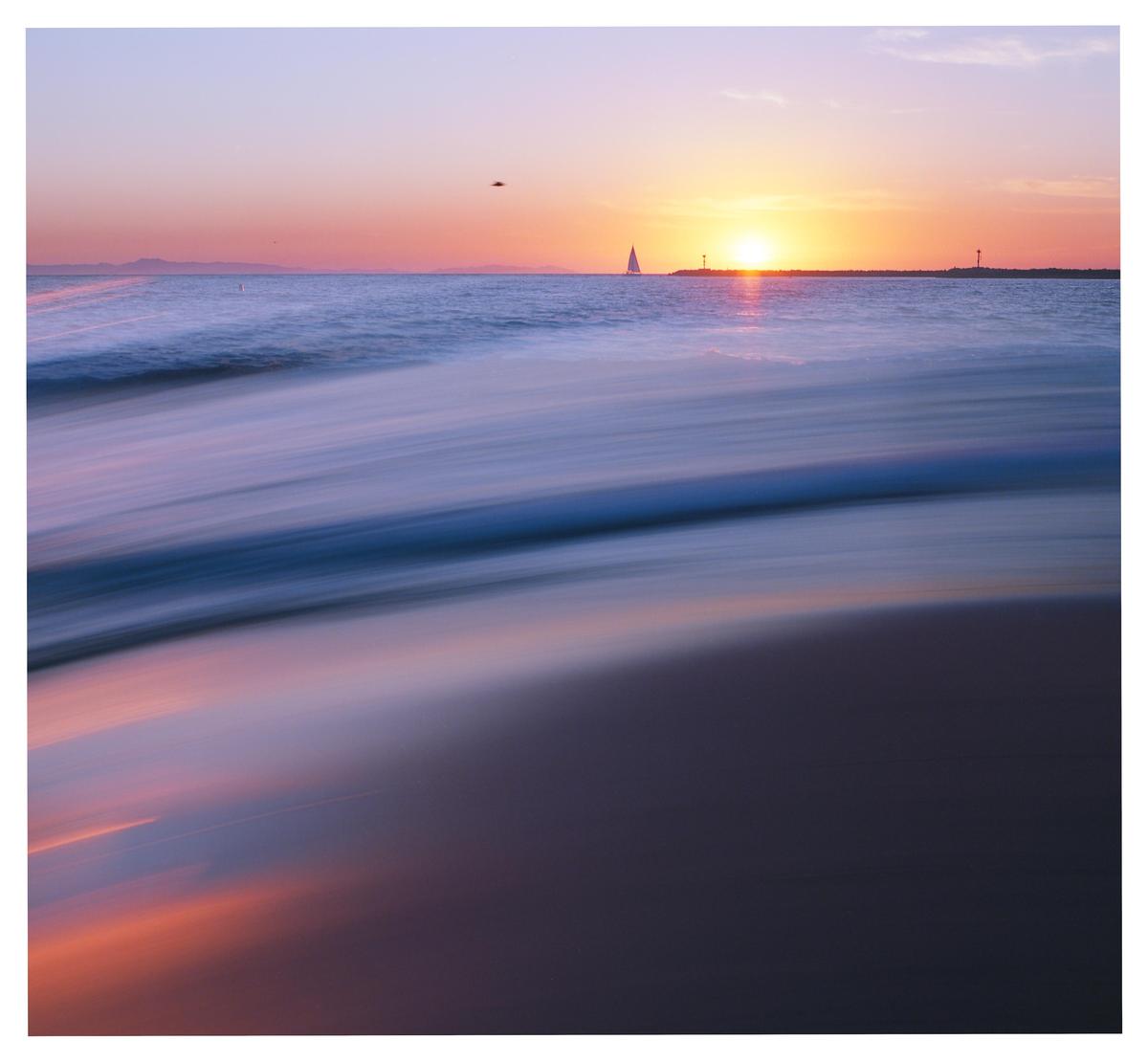 A long exposure photograph captures the motion of waves on a beach at sunset, with a sailboat and a distant landmass visible on the horizon.
