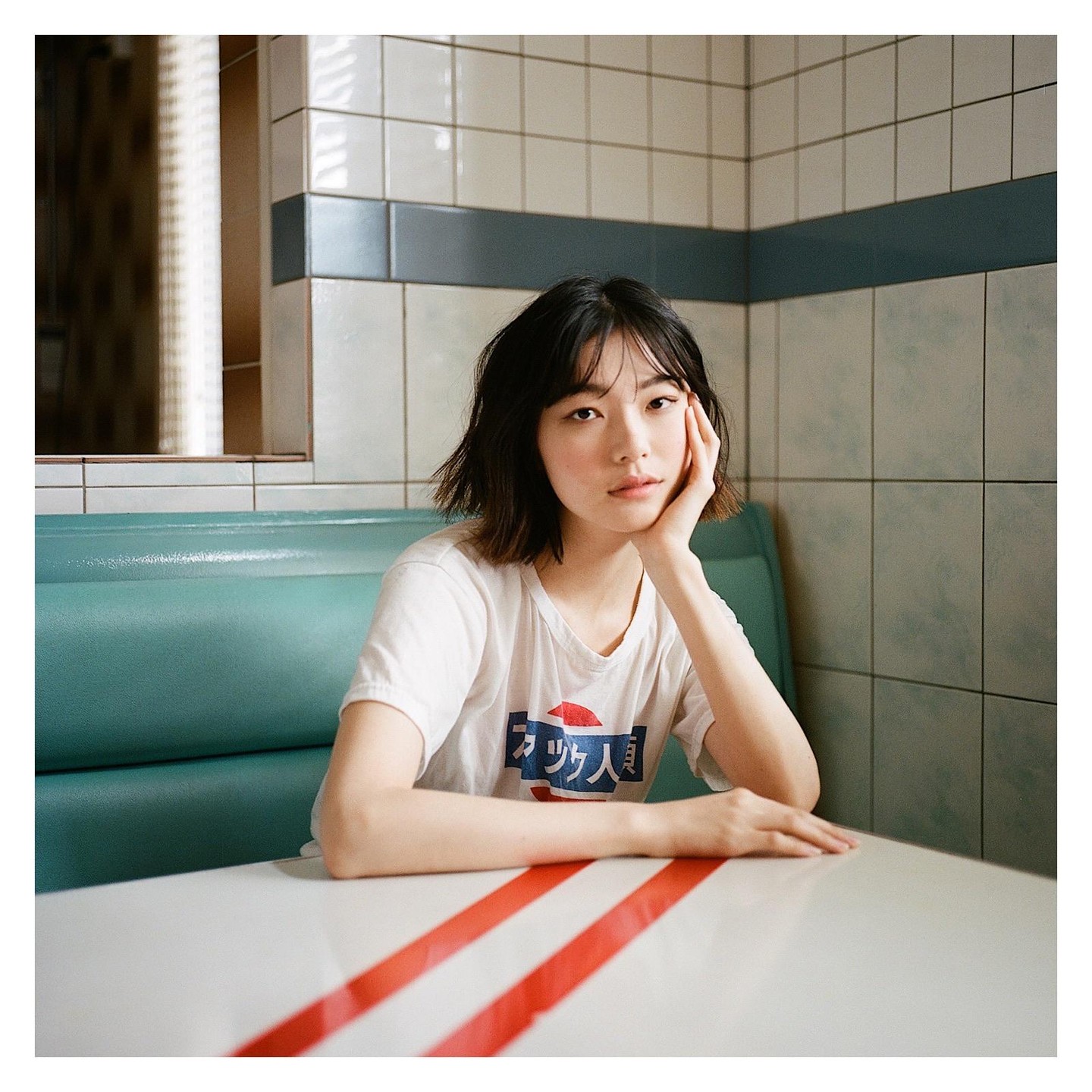 A woman with short hair sits at a diner booth with her hand on her chin.
