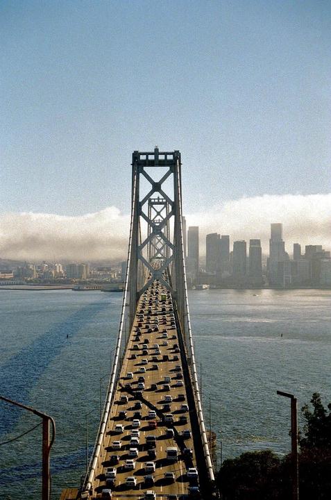A suspension bridge with heavy traffic leads to a city skyline across a body of water under a hazy sky.