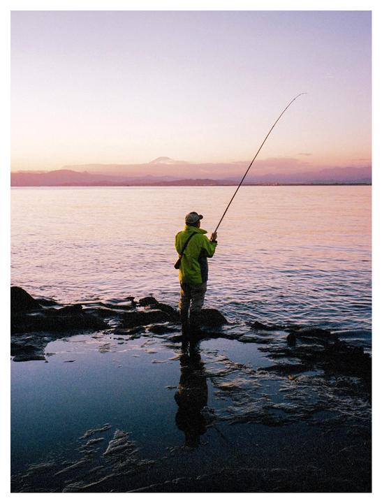 A man stands in shallow water on a rocky shore, fishing with a rod at sunset, with a mountain in the distance.