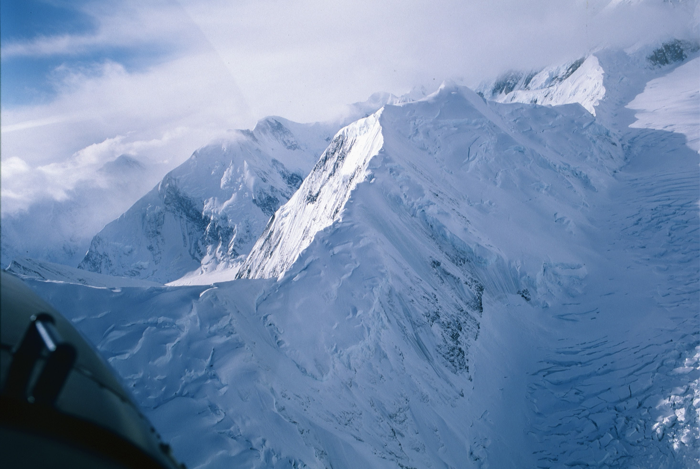 Snow-covered mountains and glaciers are visible from an aerial perspective, with a part of an aircraft in the lower left.