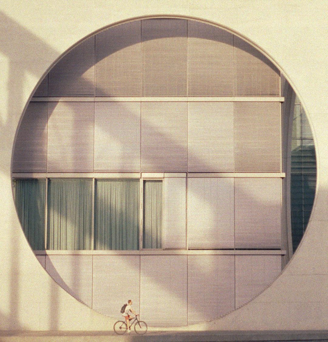 A person rides a bicycle past a building with a large circular window.
