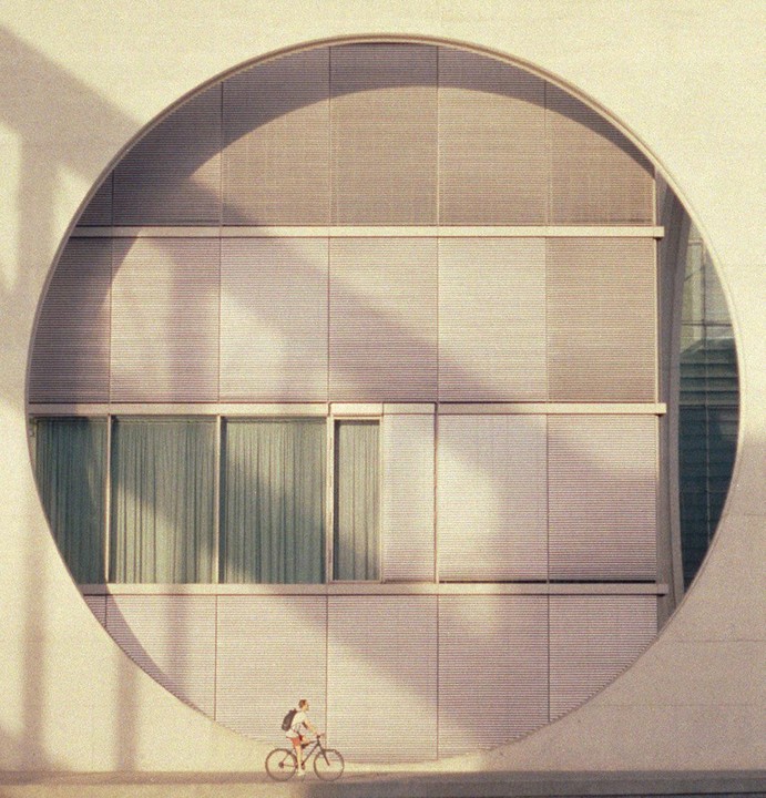 A person rides a bicycle past a building with a large circular window.