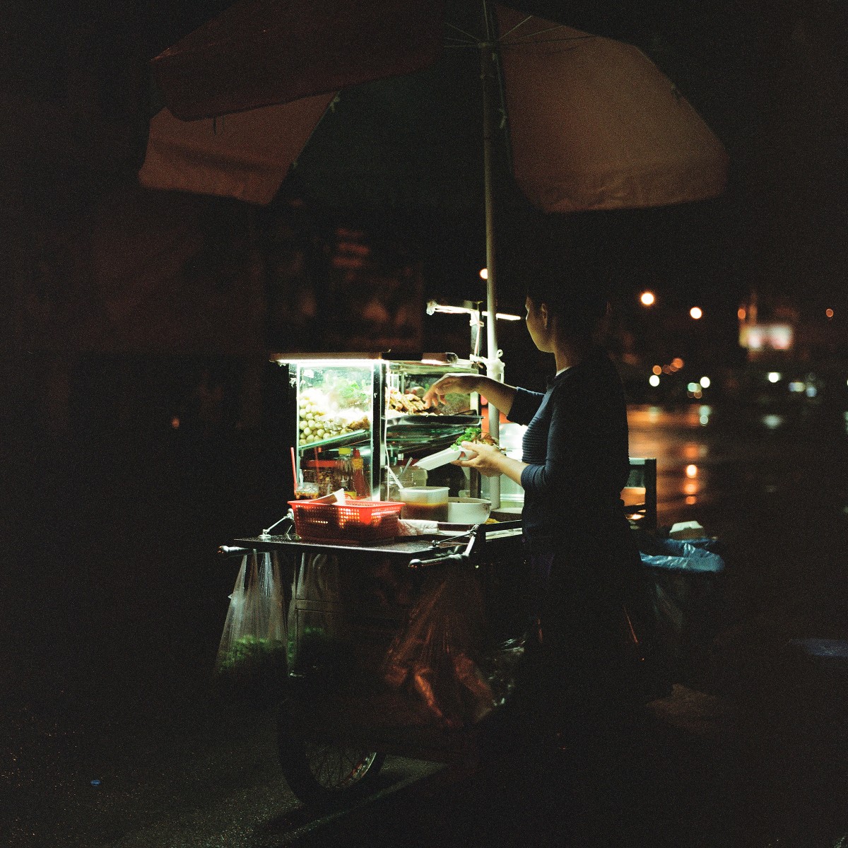 A woman stands behind a brightly lit food cart under an umbrella at night.