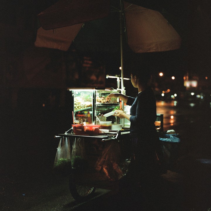 A woman stands behind a brightly lit food cart under an umbrella at night.