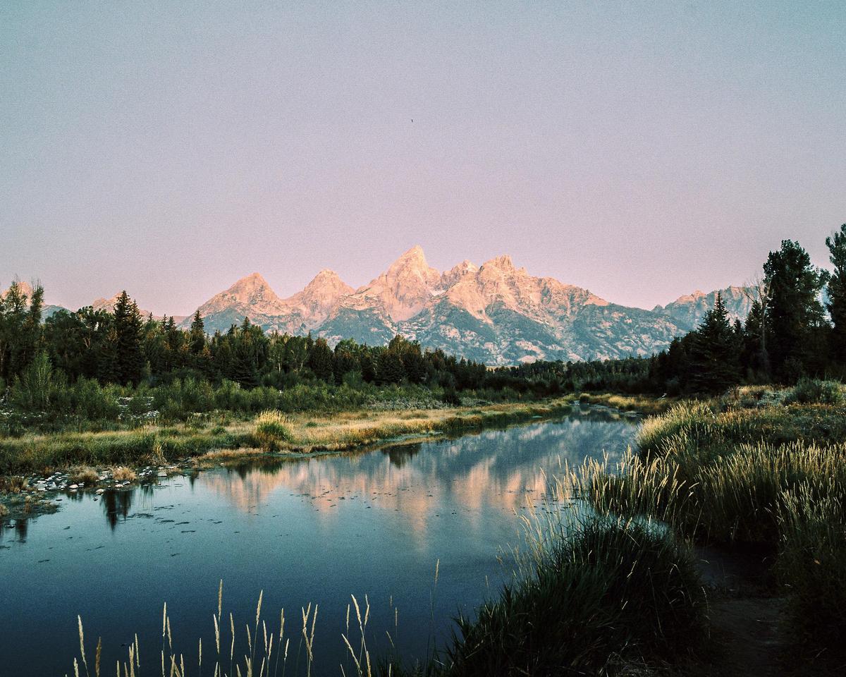 A river reflects mountains and trees under a pink sky.