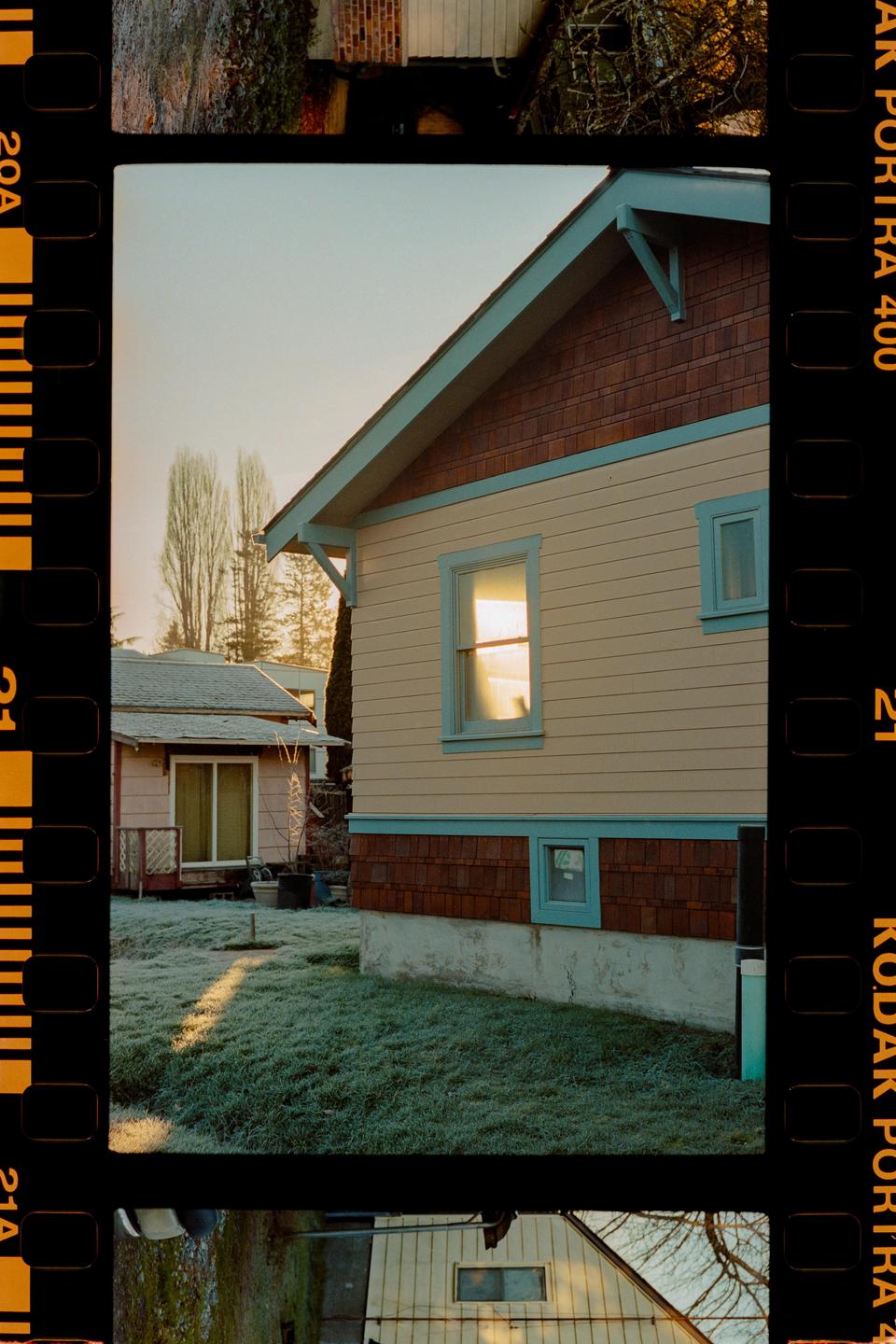 A house with light yellow siding and blue trim stands next to a smaller pink house, with frost covering the grass in the foreground.