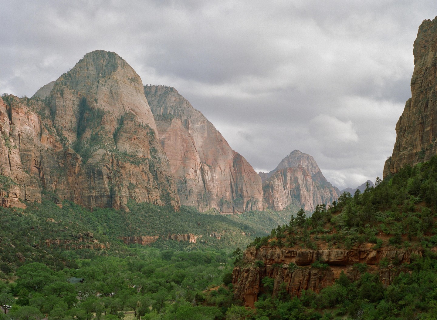 A valley filled with green trees is surrounded by tall, rocky mountains under a cloudy sky.