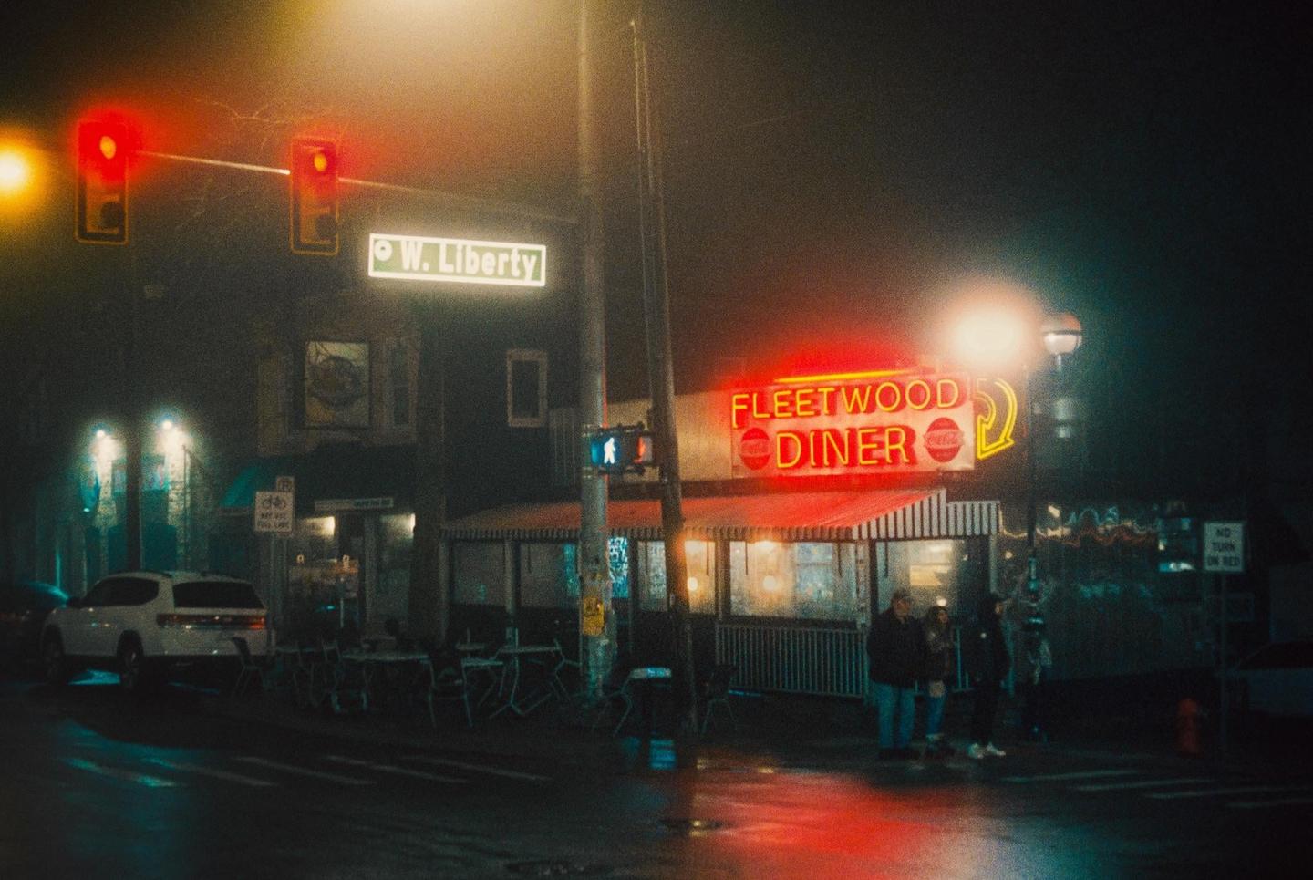 A diner with a bright neon sign is visible at night on a foggy street corner with traffic lights and a street sign.