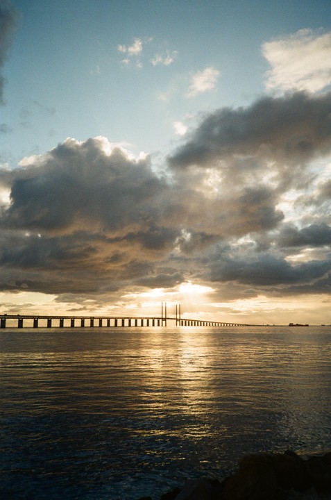 The Øresund Bridge stretches across the water under a dramatic, cloudy sky with the sun setting behind it.