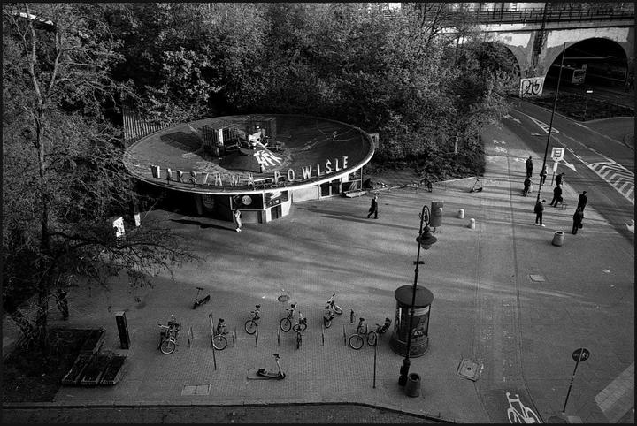 A high-angle view shows a circular building with "Powisle" written on its roof, surrounded by trees, a street, and several people.