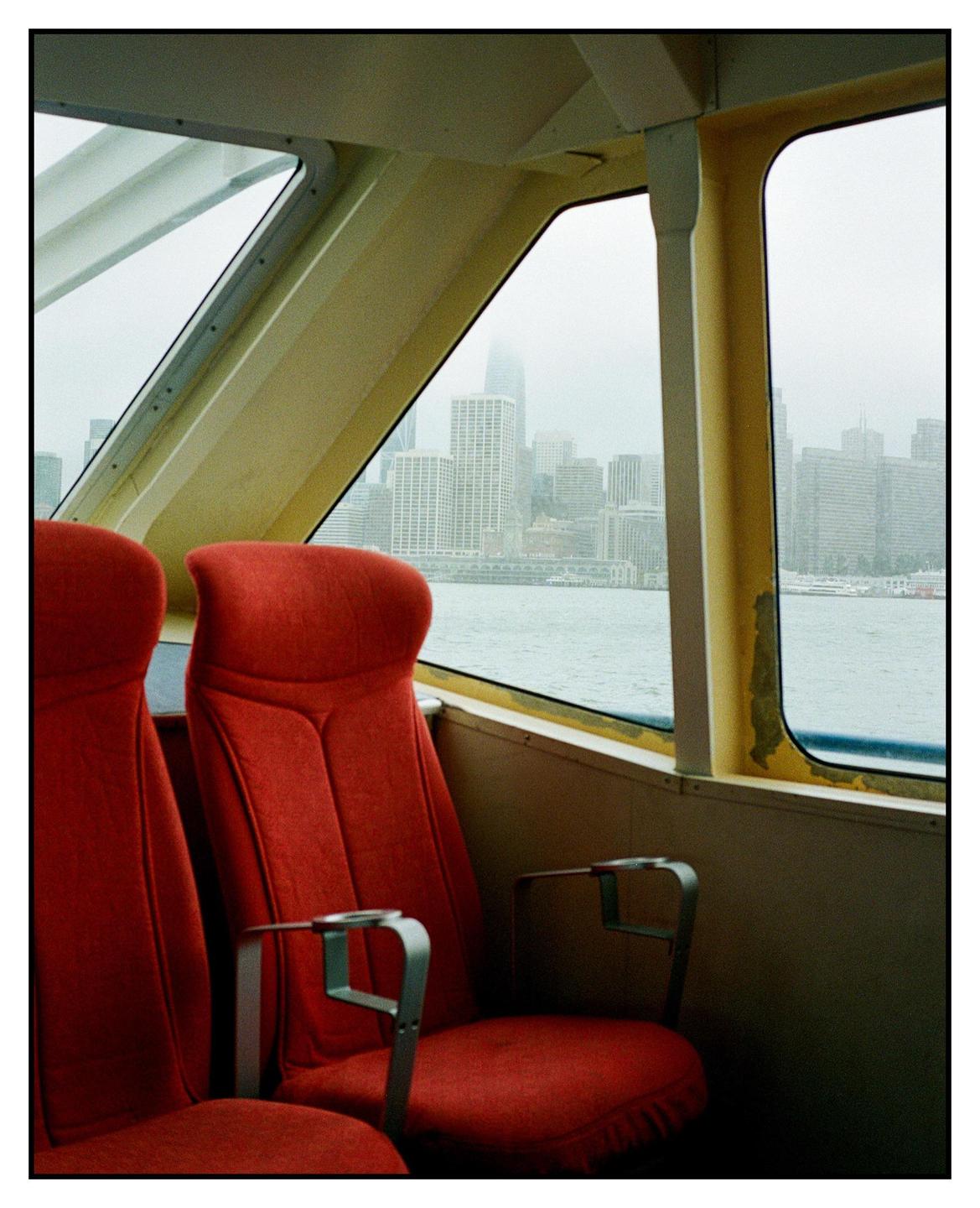 Two red seats are visible inside a ferry with a city skyline in the background.