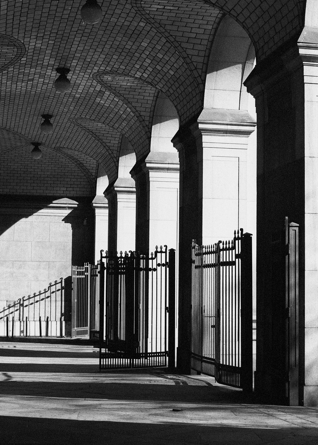 A series of arches with a brick ceiling and ornate metal gates are illuminated by strong sunlight, creating deep shadows.