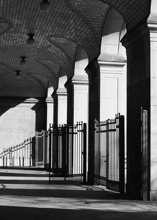 A series of arches with a brick ceiling and ornate metal gates are illuminated by strong sunlight, creating deep shadows.