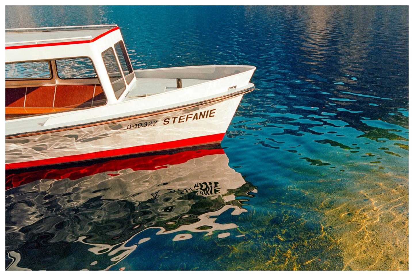 A white boat with a red stripe is reflected in clear blue water next to a sandy bottom.