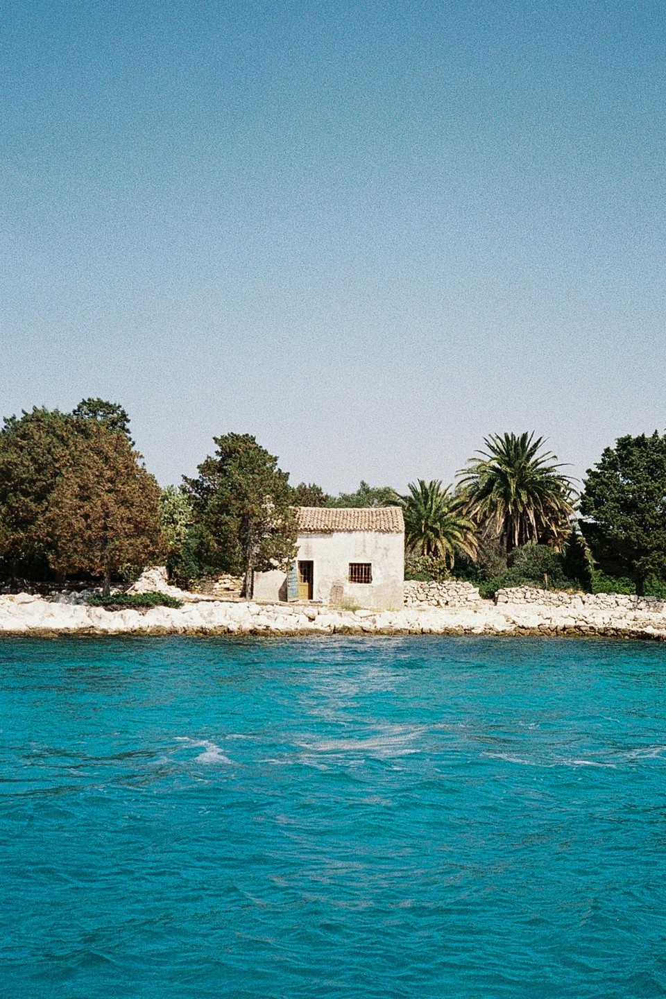 A small house with a tiled roof sits on a rocky shore next to turquoise water, surrounded by trees and palm trees.