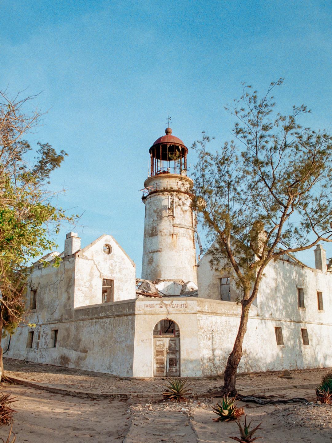 An old, weathered lighthouse stands among trees and sandy ground under a clear sky.