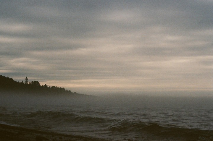 A body of water with waves crashes against a tree-lined shore under a cloudy sky.